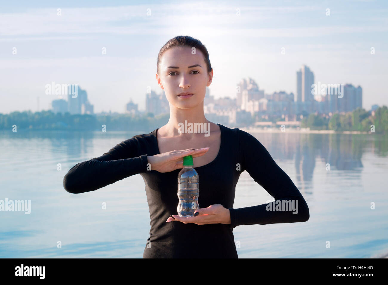 Young pretty fitness woman holds water bottle at morning river and city on backround Stock Photo