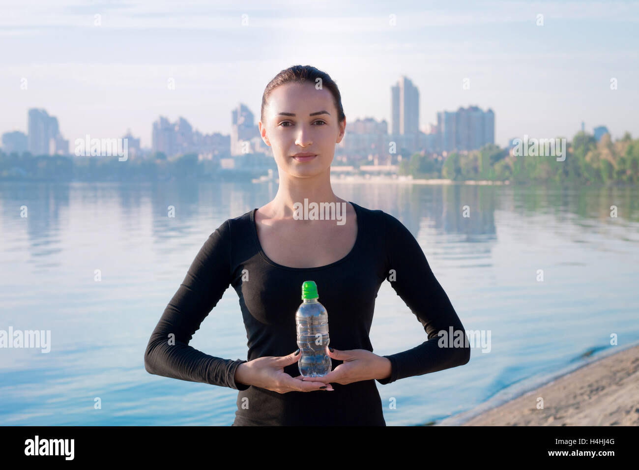 Young pretty fitness woman holds water bottle at morning river and city on backround Stock Photo