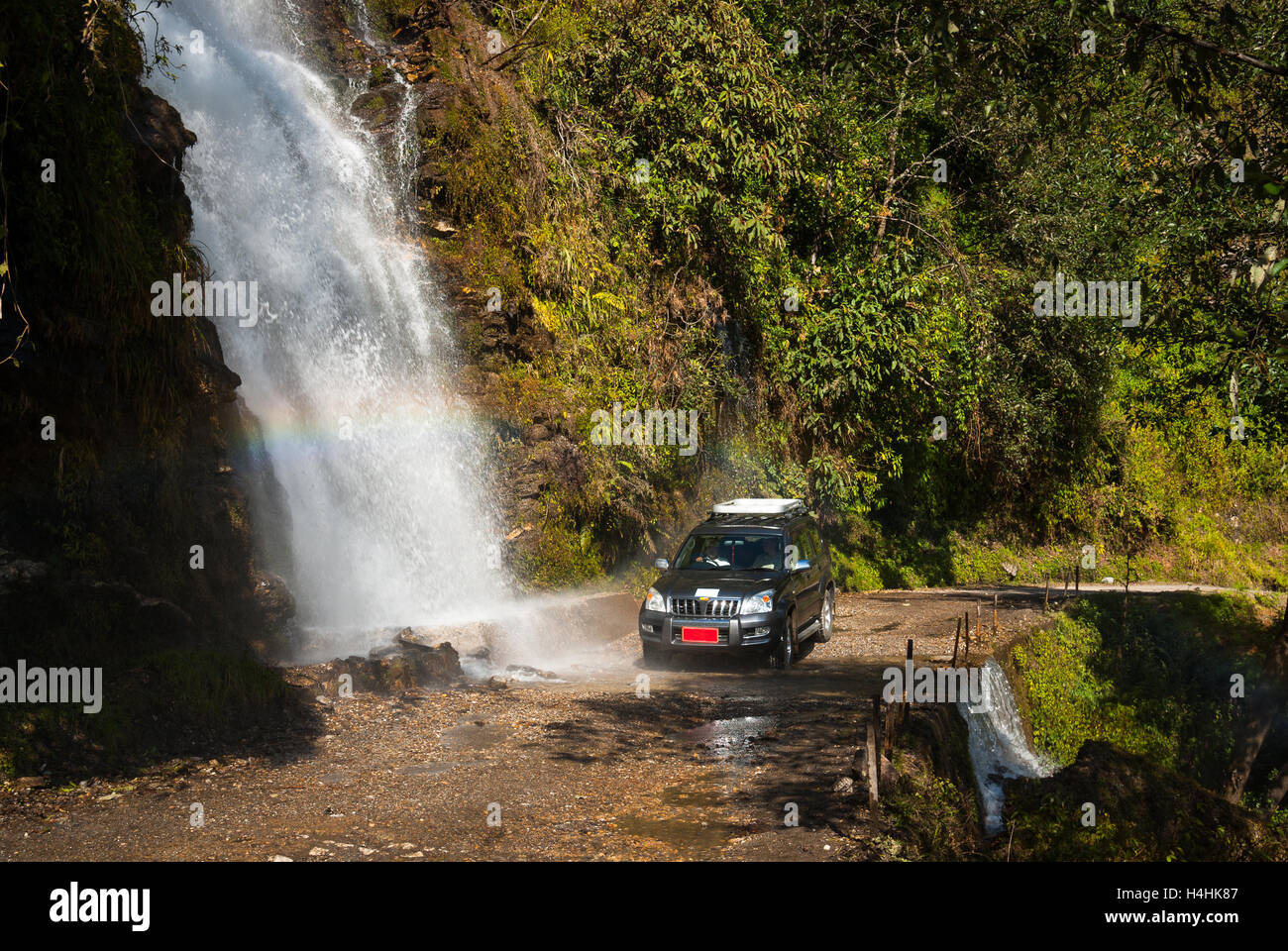Four-wheel drive vehicle driving through waterfall over road south of Trongsa, Bhutan. A rainbow is visible in the mist. Stock Photo