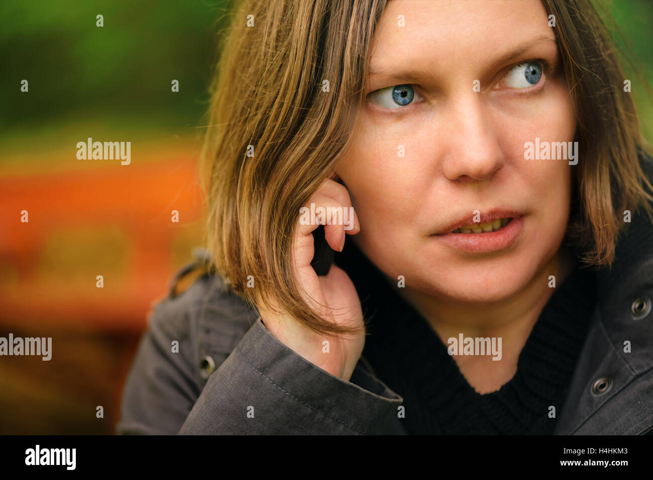 Woman with serious expression talking on mobile phone in park, dramatic light coming through treetops and falling on her face Stock Photo