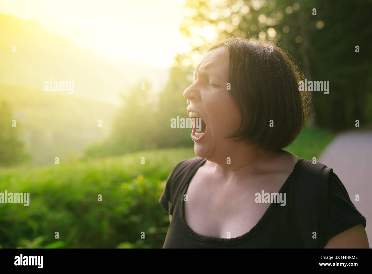 Young adult woman yawning outdoors in the morning, sleepy tired female person walking in nature Stock Photo