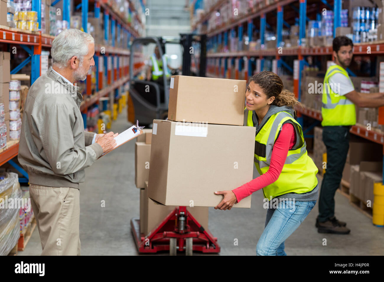 Warehouse manager noting on clipboard while female worker carrying cardboard boxes Stock Photo