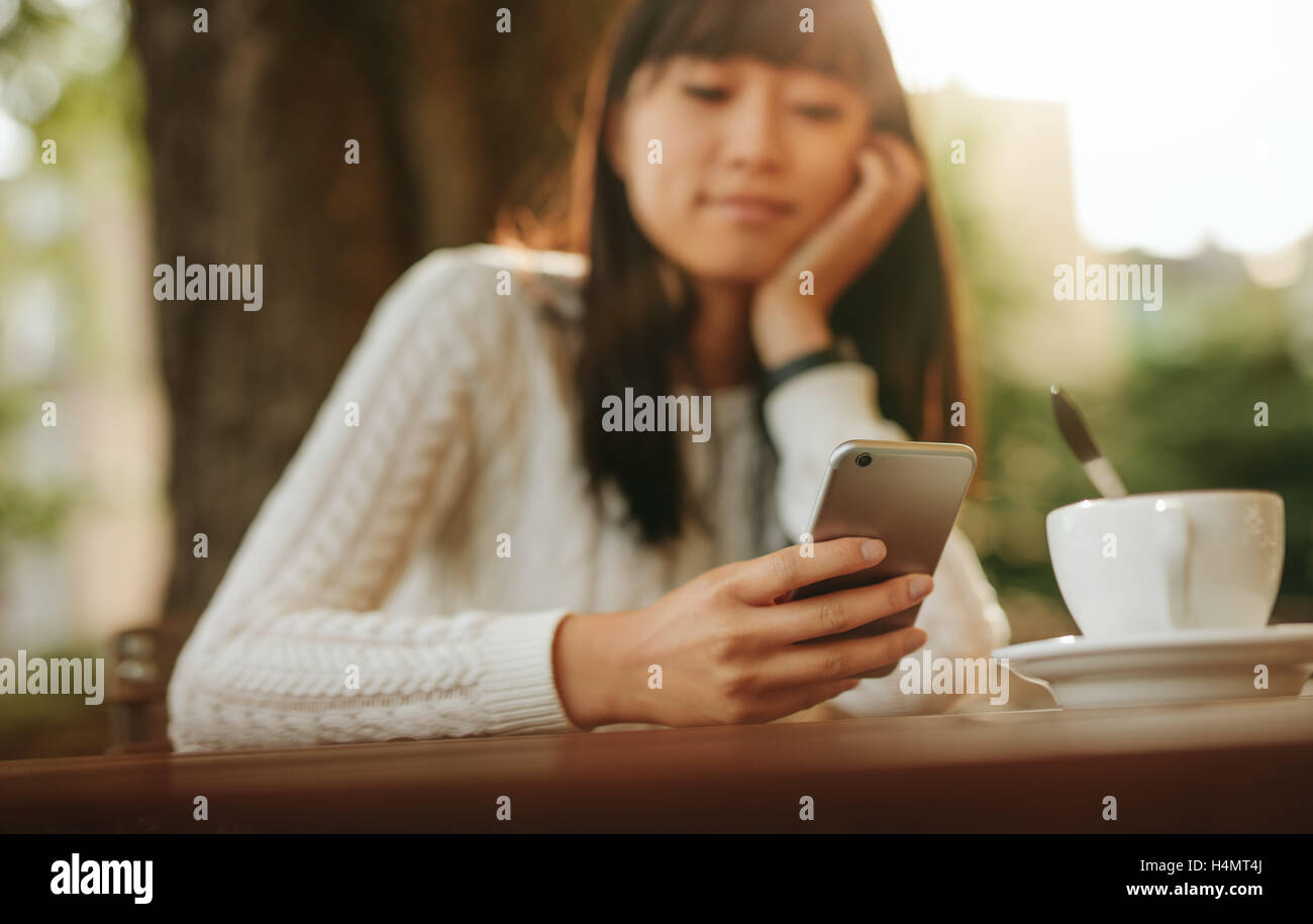 Young chinese woman looking at mobile phone in her hand while sitting at outdoor cafe. Asian female model reading text message o Stock Photo