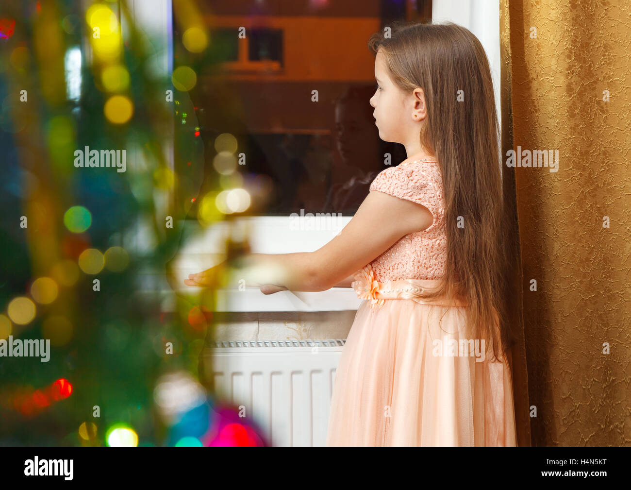 little girl with long brown hair and in pink dress standing at the window at Christmas Stock Photo