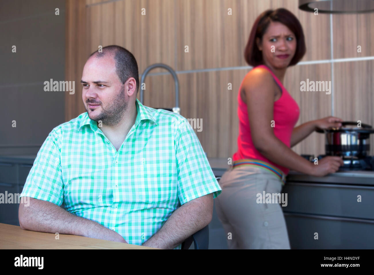 overweight man is sitting at kitchen table and hispanic woman is cooking and looking angry Stock Photo