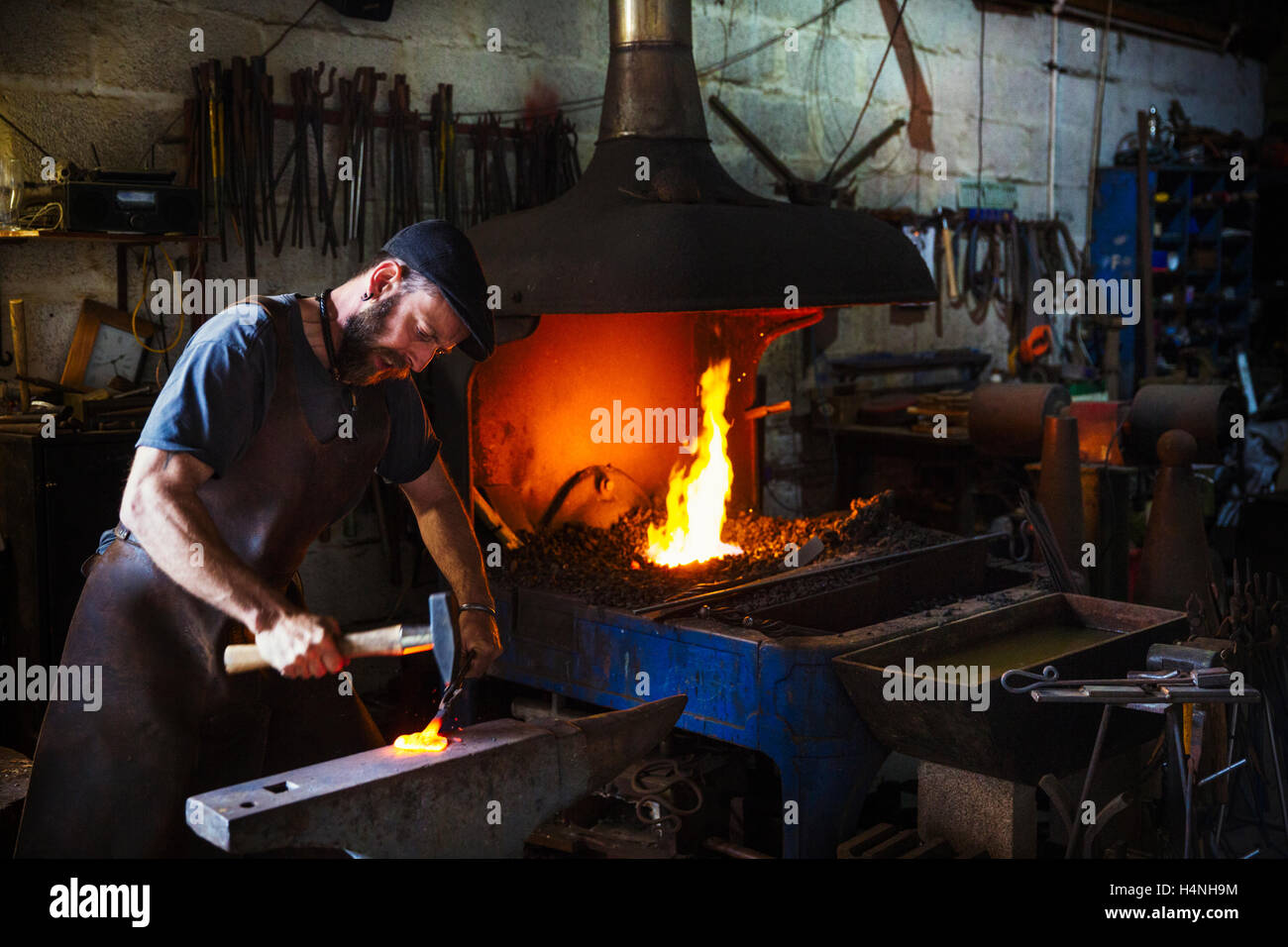 A blacksmith strikes a piece of red hot metal on anvil with a hammer in a workshop. Stock Photo