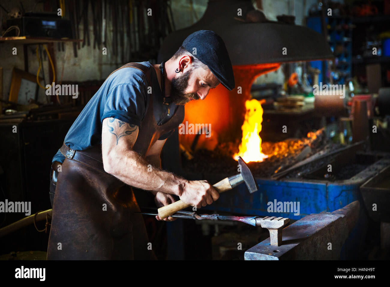 A blacksmith uses complex tools to hammer a cone of red hot metal on anvil in a workshop. Stock Photo