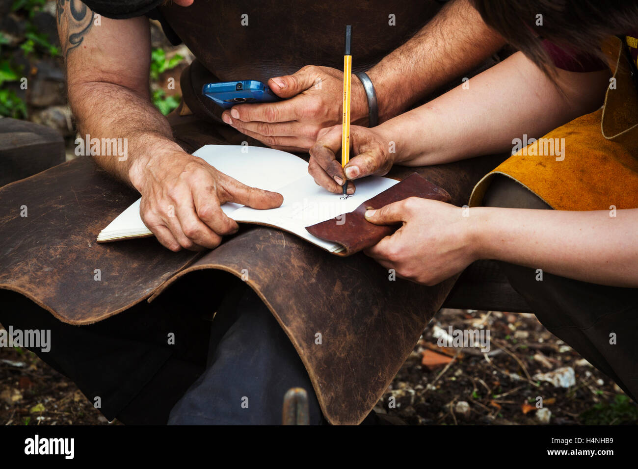 Man and woman, blacksmiths wearing aprons writing into a notebook sat in a garden. Stock Photo