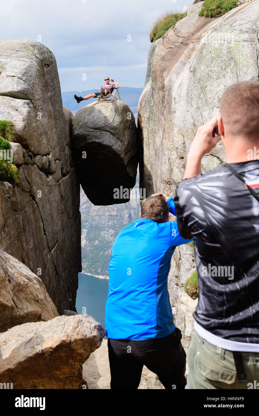 Tourists taking photos of friend, sitting dangerously in a beach chair with a can of beer on Kjeragbolten, a rock hanging 1100 meters above the fjord Lysefjorden, Norway Stock Photo
