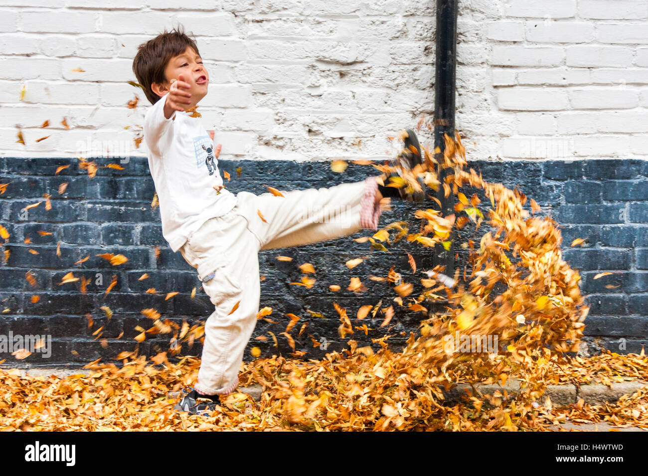 Caucasian child, boy, seven years old, kicking brown autumn leaves in street by white wall. Side view. Stock Photo