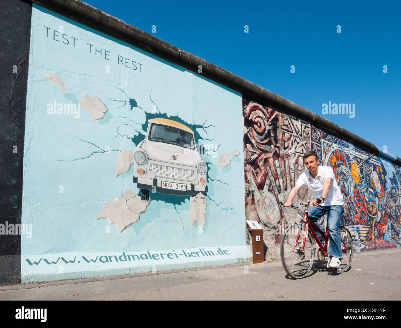 Man cycles past painting of Trabant car on wall at East Side Gallery at former Berlin Wall in Friedrichshain/Kreuzberg in Berlin Stock Photo