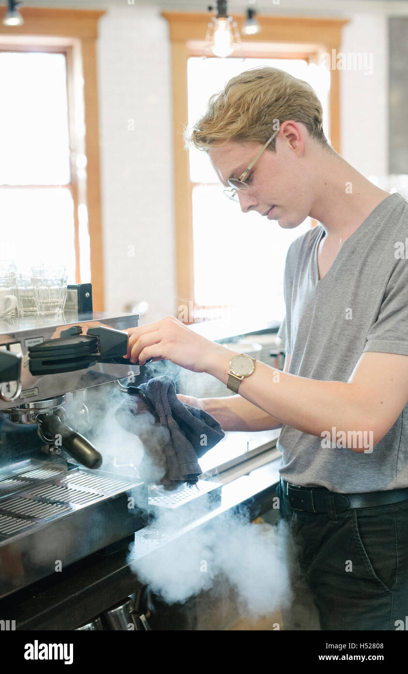 Young blond man with spectacled in front of espresso machine, steam coming out of machine. Stock Photo