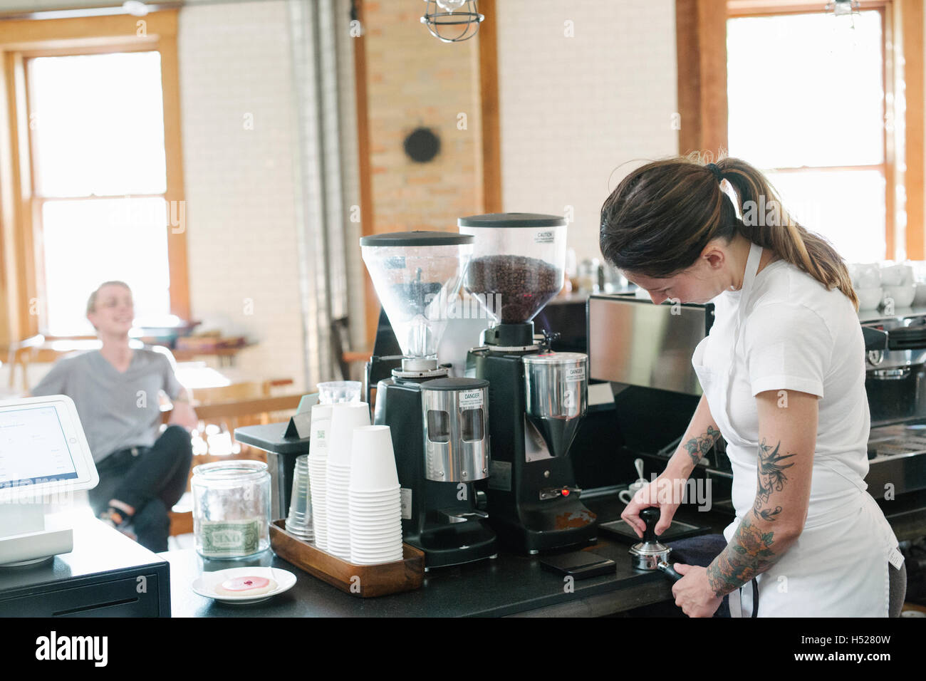 Woman wearing a white apron, in front of espresso machine, holding a portafilter. Stock Photo