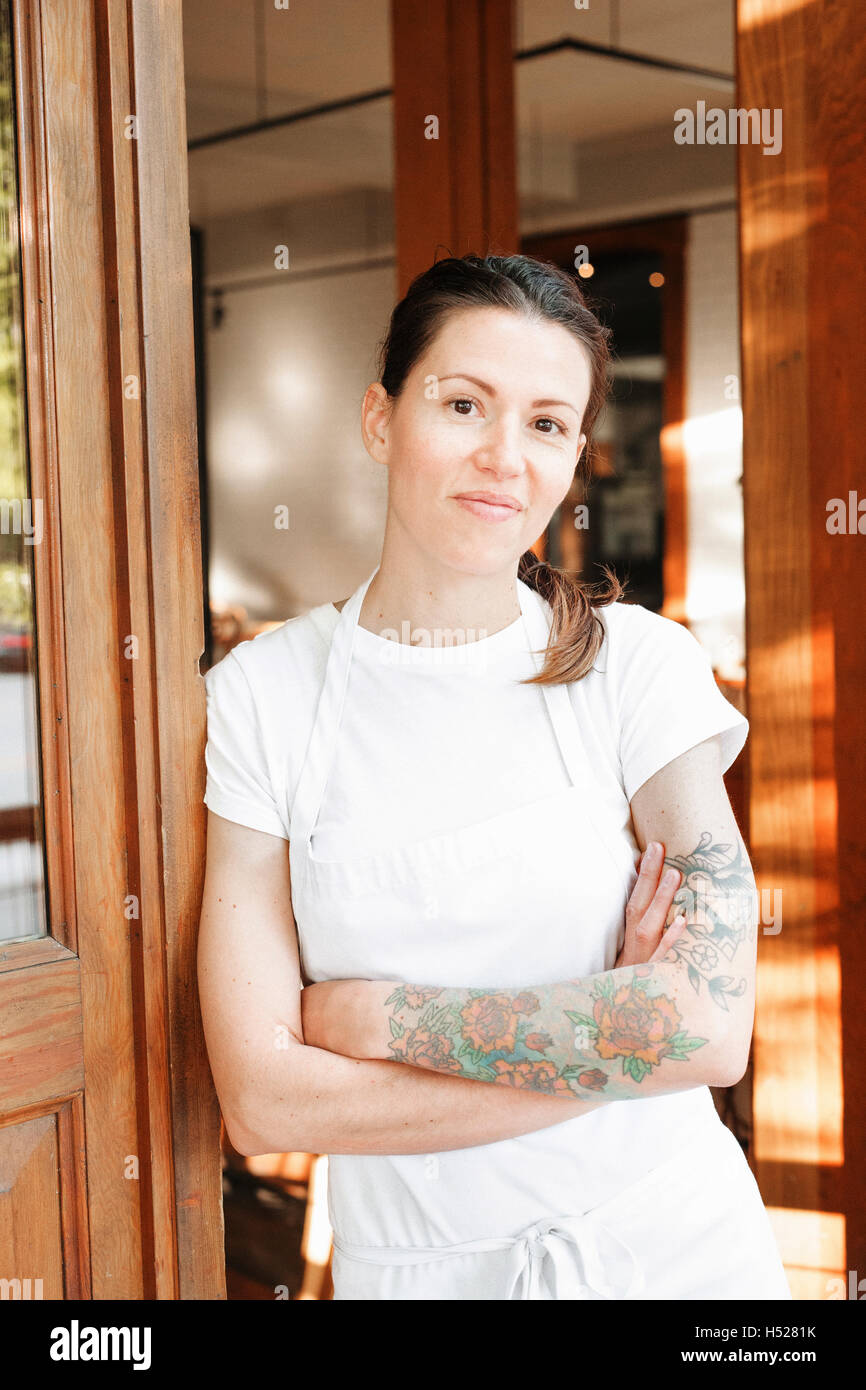 Portrait of a woman wearing a white apron with her arms crossed in a doorway, tattoos on her left arm. Stock Photo