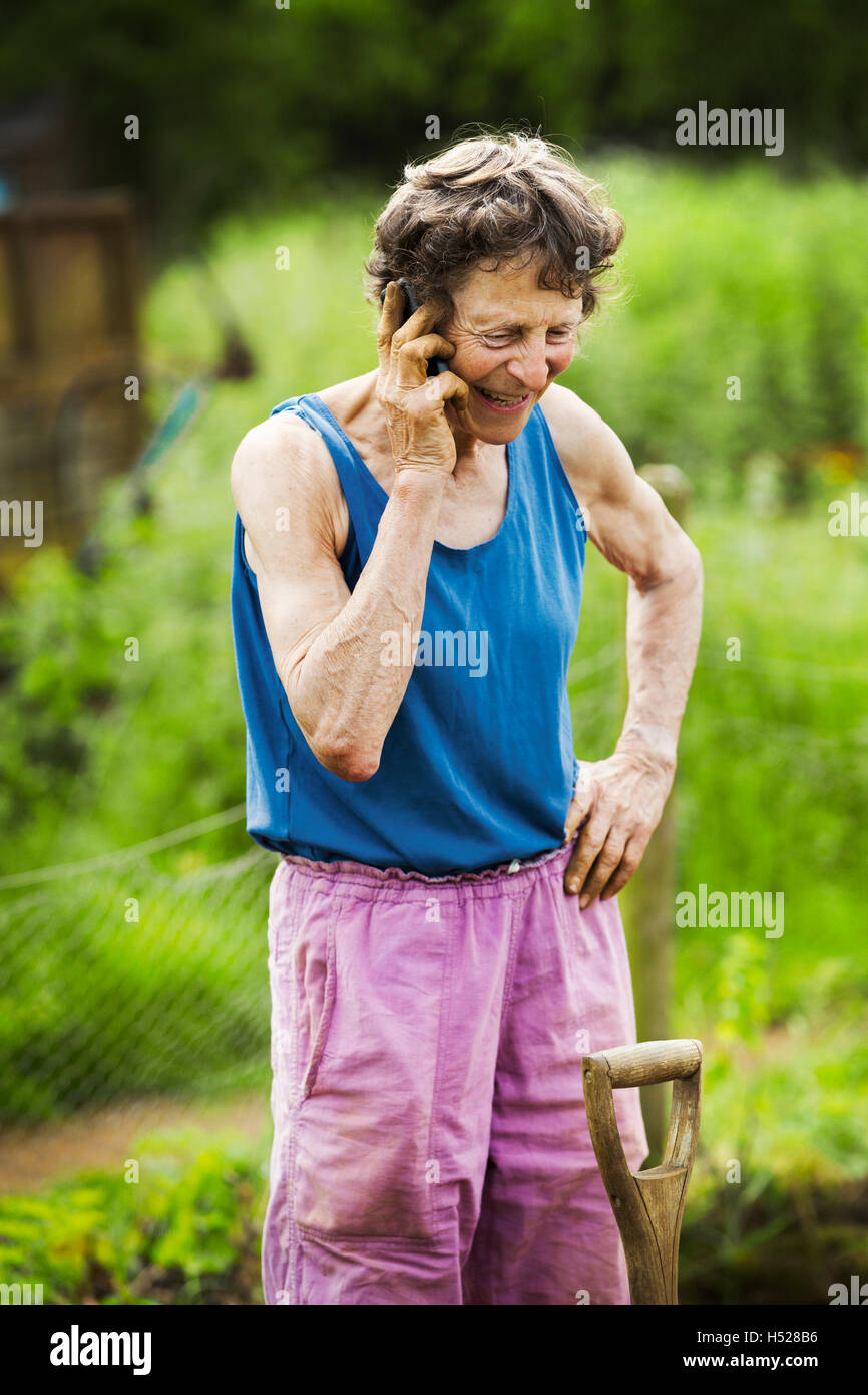 A woman talking on the phone with a shovel by her leg in a farm. Stock Photo