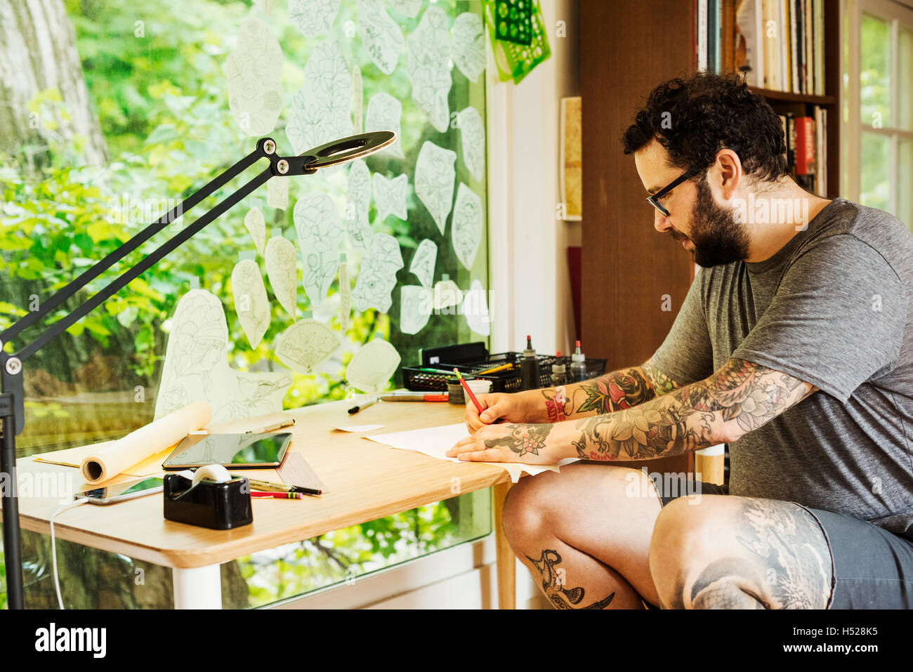 Bearded man with tattoos on his arms and legs sitting at a desk, drawing. Stock Photo