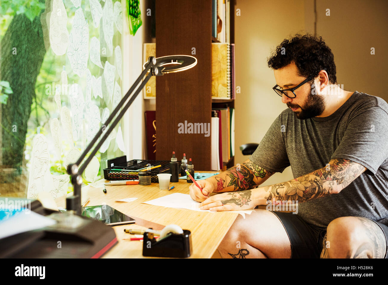 Bearded man with tattoos wearing glasses, sitting at a desk, drawing. Stock Photo