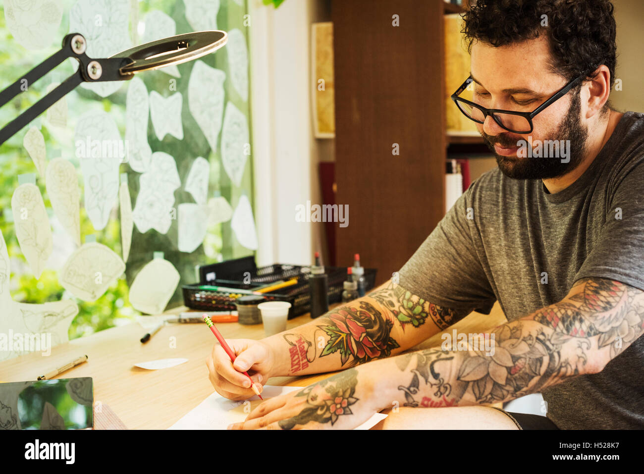 Bearded man with tattoos on his arms, sitting at a desk, drawing. Stock Photo