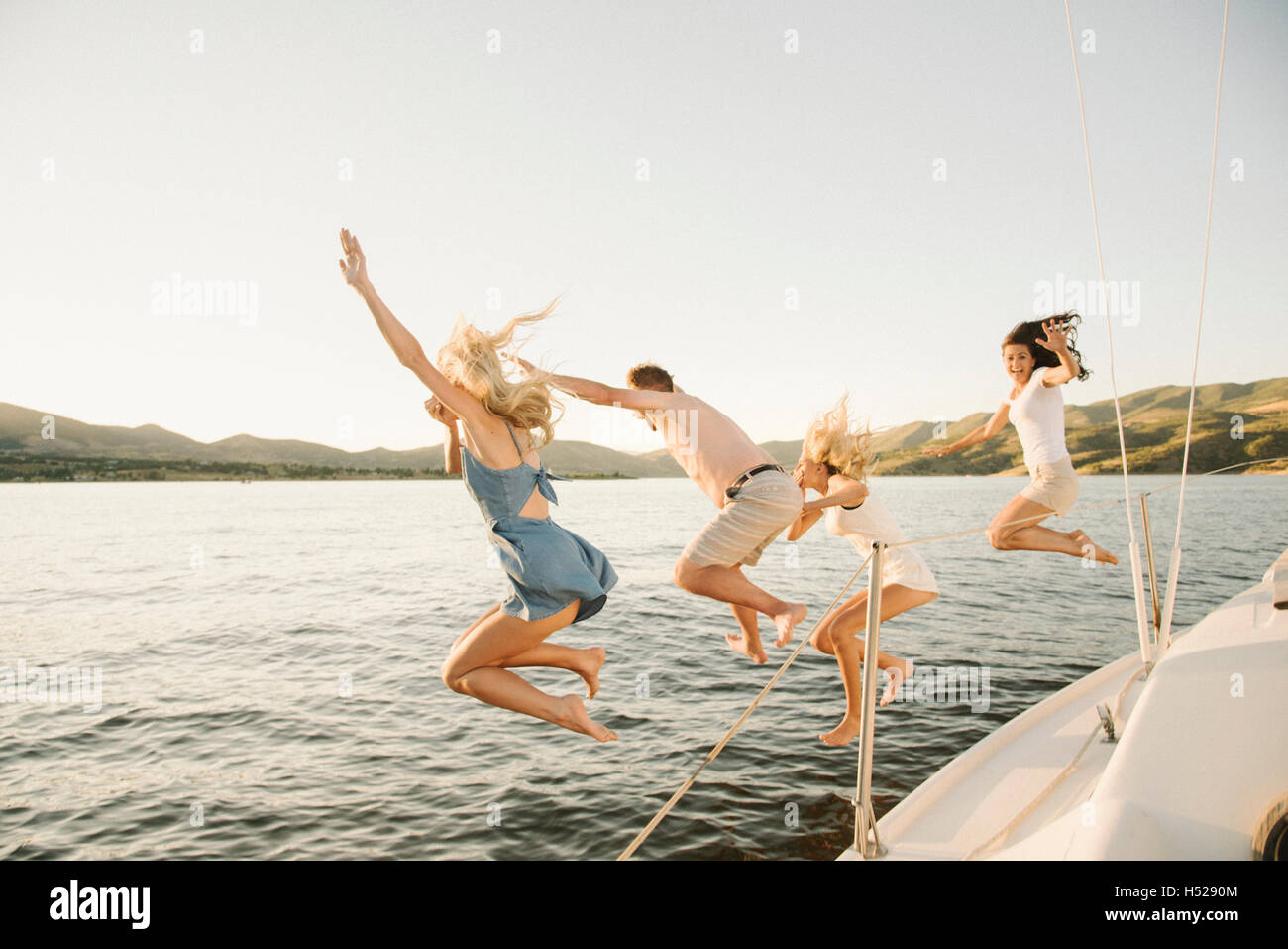 Four people jumping off the side of a sail boat into a lake. Stock Photo