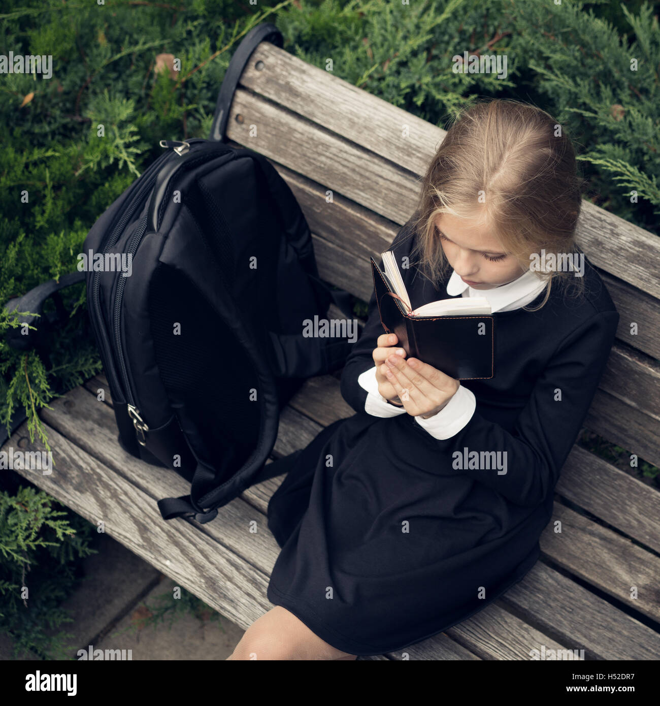 Beautiful blond girl in a school uniform sits on park bench and reading book. Stock Photo
