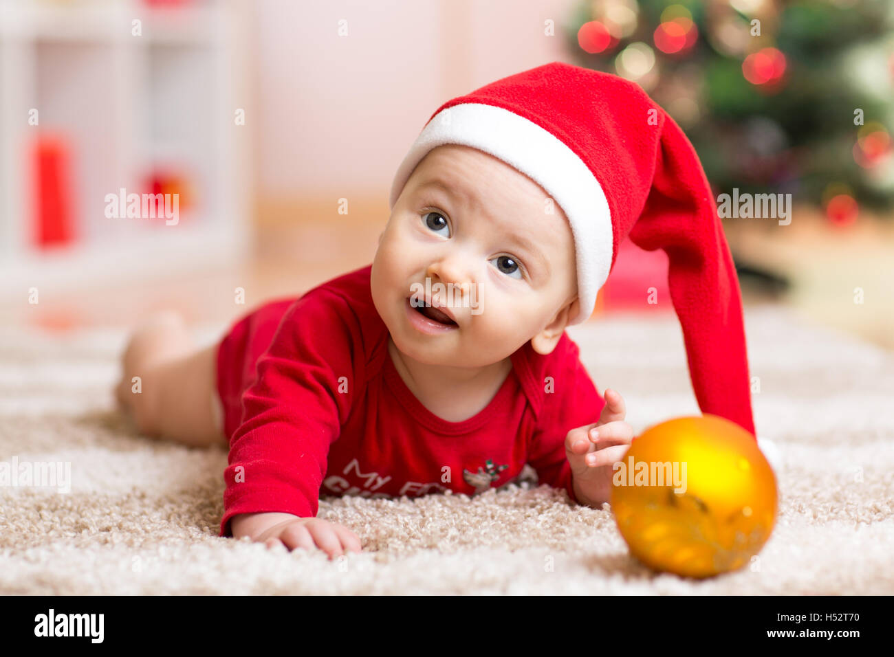 Funny baby wearing Santa hat and suit. Kid boy lying on tummy in front of Christmas tree at home Stock Photo