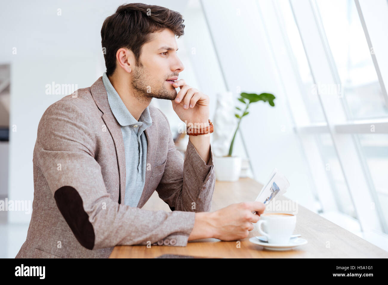 Thoughtful young businessman holding newspaper while having coffee break indoors Stock Photo