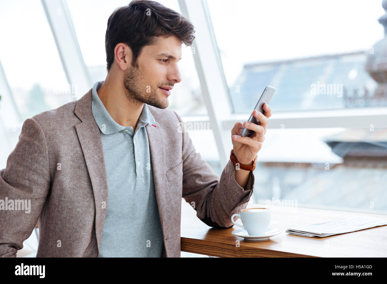 Young casual man in jacket texting message on smartphone while sitting indoors Stock Photo