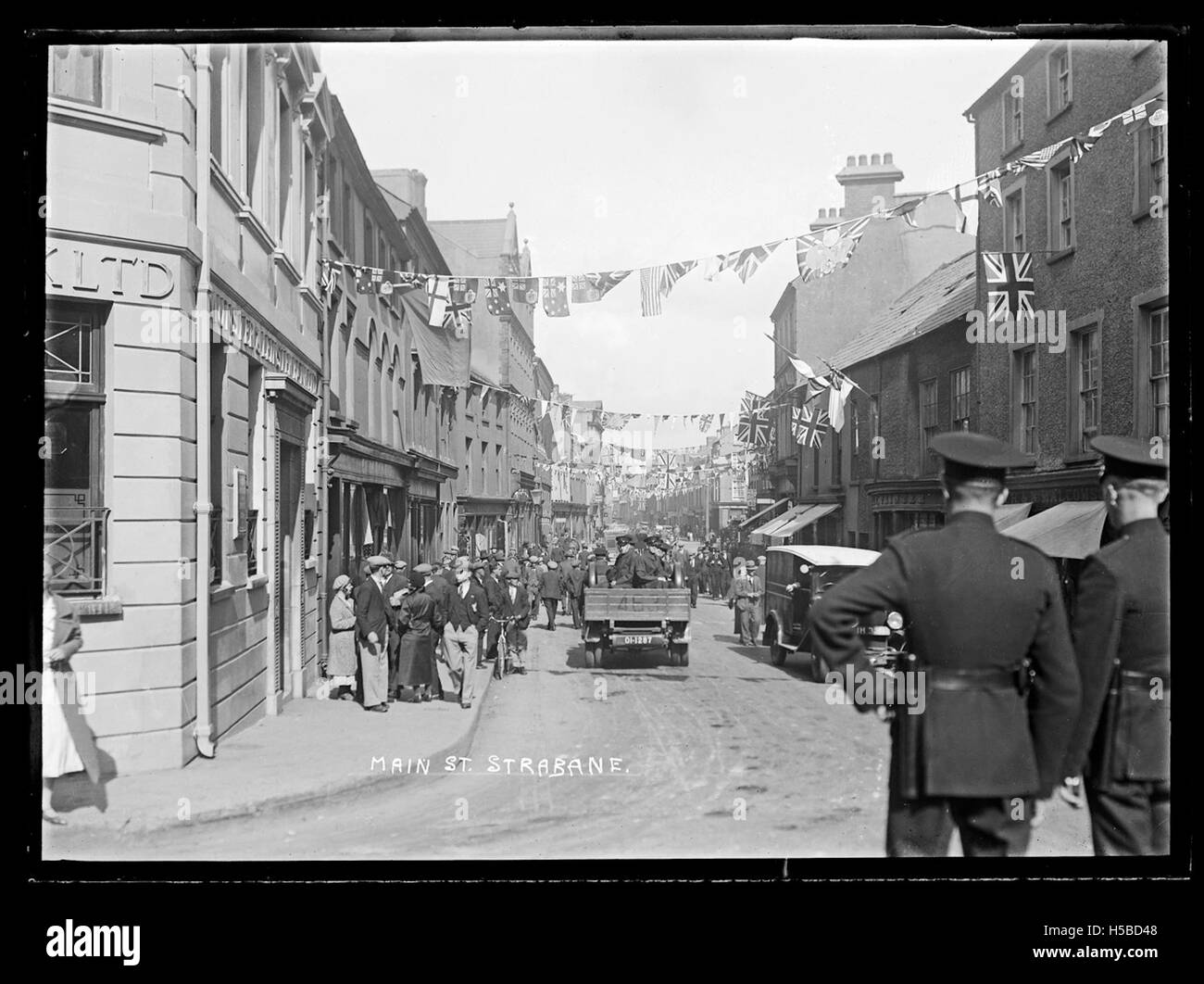 A view down a bustling street in Strabane town centre, Northern Ireland, with cars parked along the curb, pedestrians walking, and traditional architecture lining both sides of the road.
