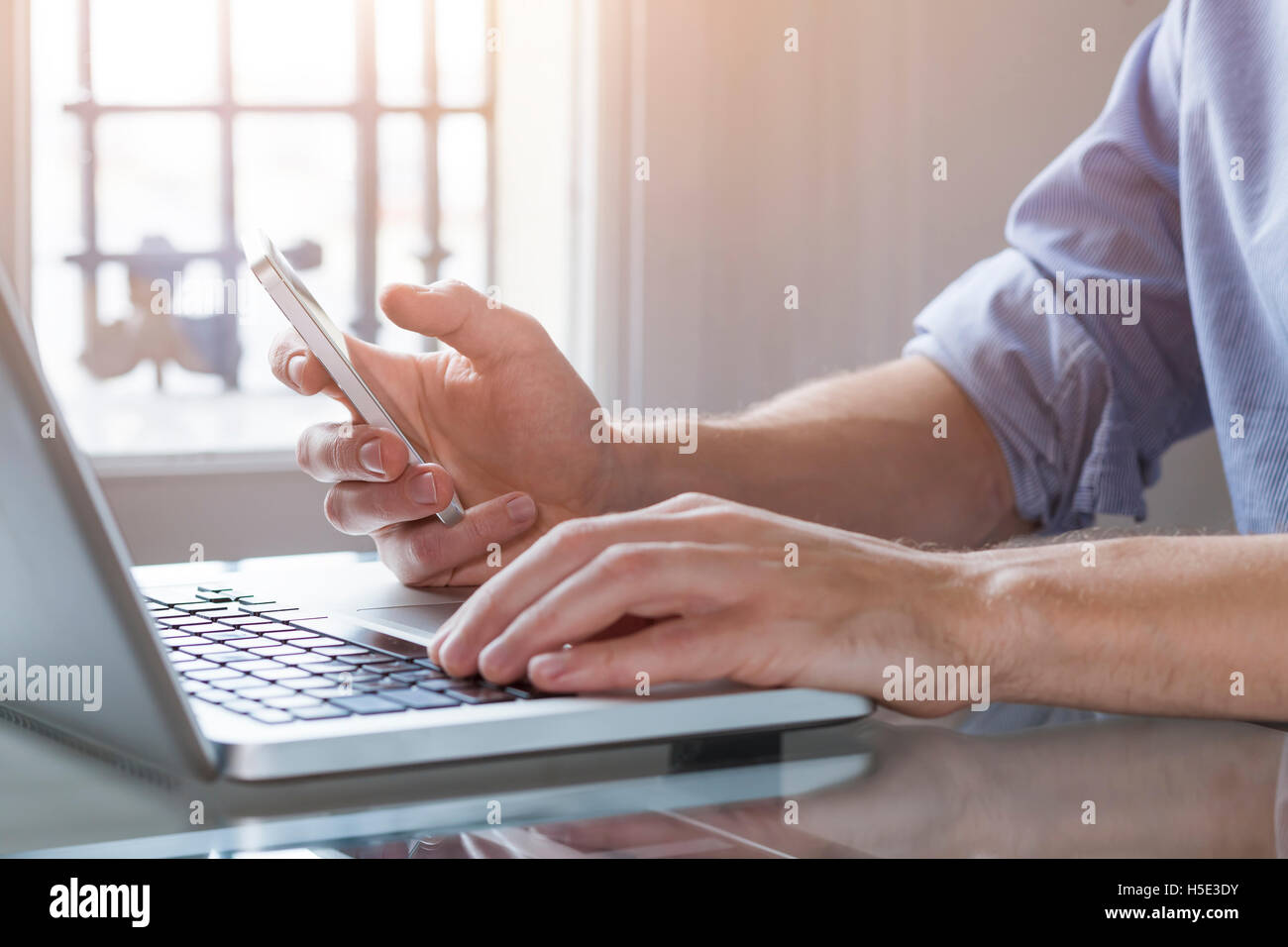 Young man working from home with laptop computer and smart phone. Close-up of hands with day light in background Stock Photo