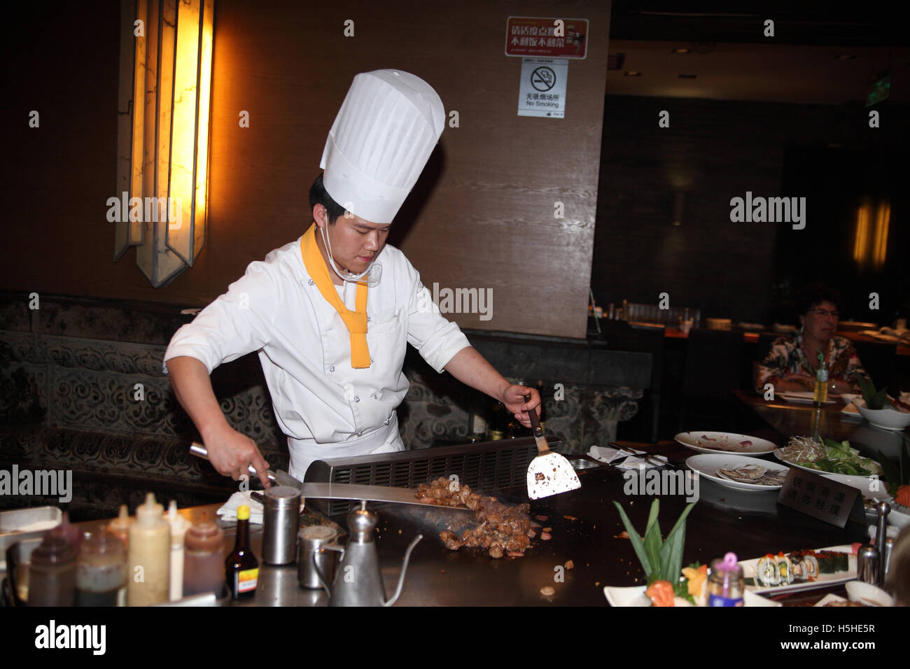 Chinese cook in a Chinese restaurant, wearing white uniform and a cap, stir fries pieces of meat in front of the diners. Beijing, China. Stock Photo