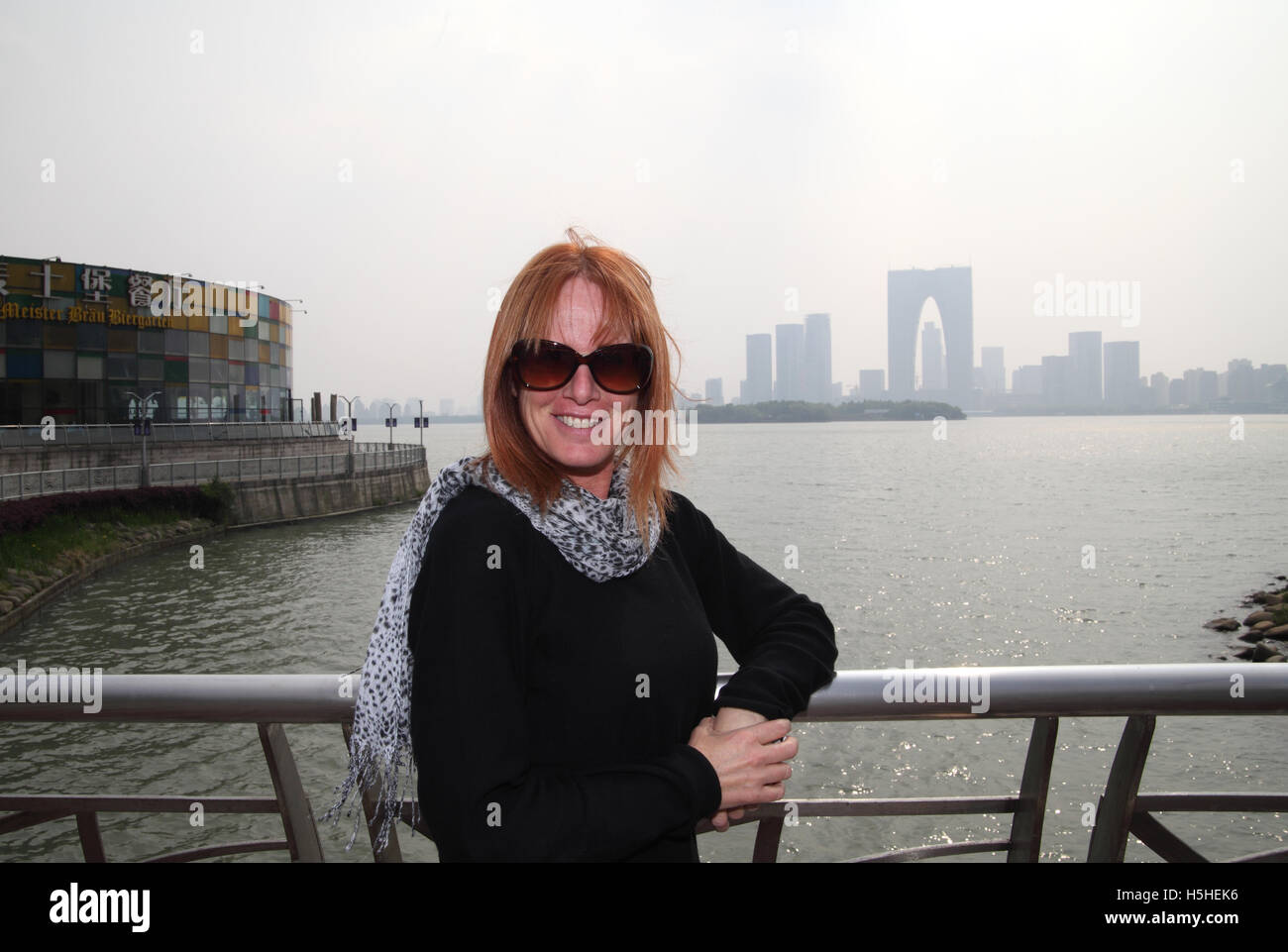 A red hair Israeli tourist smiles to the camera with the Jinji Lake, the Oriental Gate and other high rise buildings behind her. Stock Photo