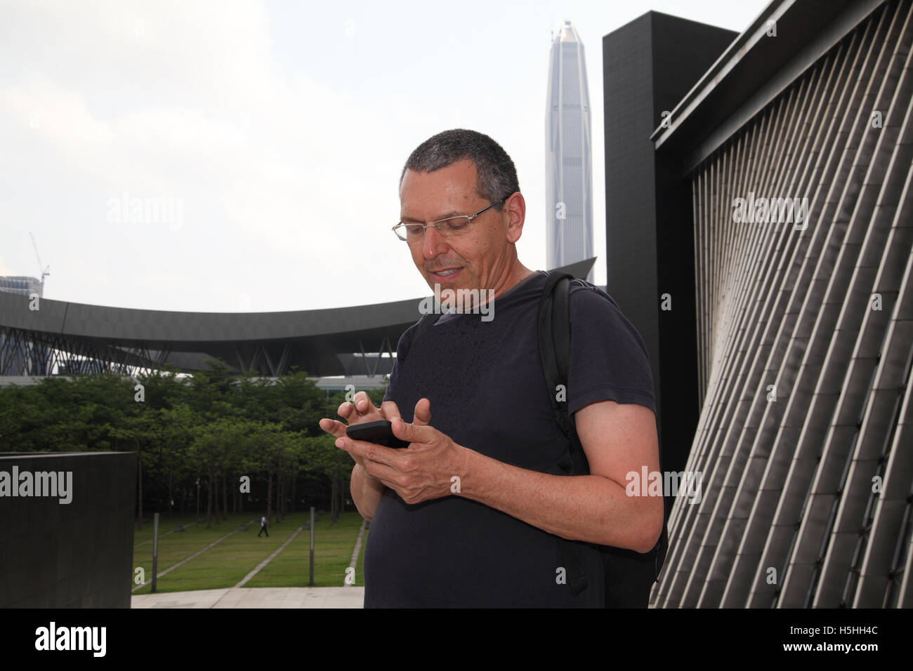 An Israeli tourist smiles while text messaging as he stands near the wall of the Shenzhen Library with the PIng An IFC behind Stock Photo