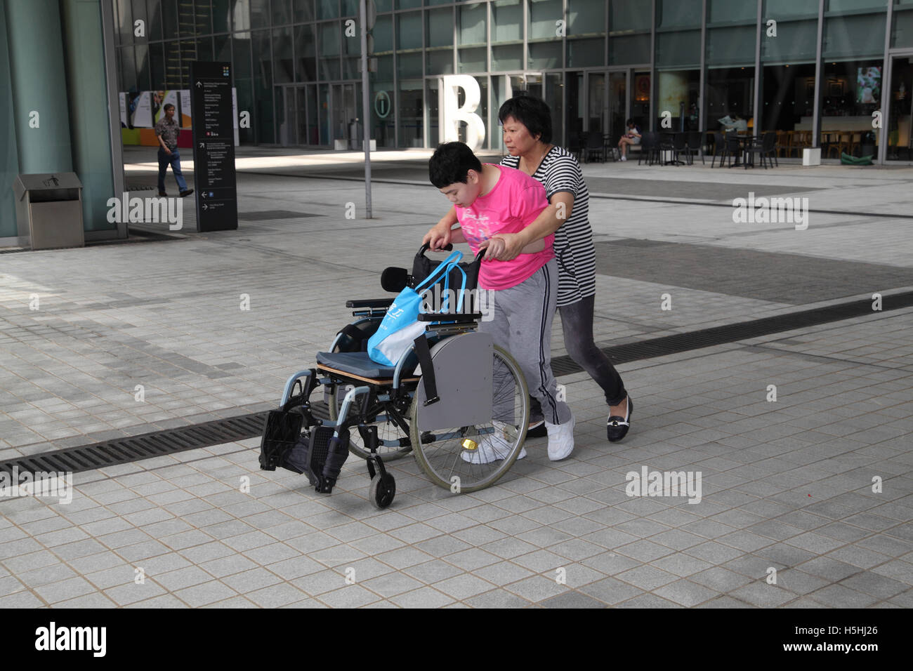 A Chinese mother is making her 20 year old brain injured daughter exercise by helping her push and walk behind the wheelchair Stock Photo