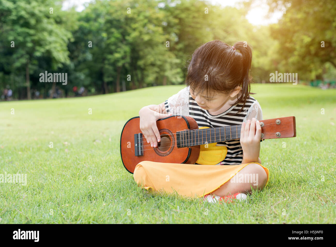 Asian little kids girl sitting on grass and play ukulele in park. Kids funny playing ukulele Stock Photo