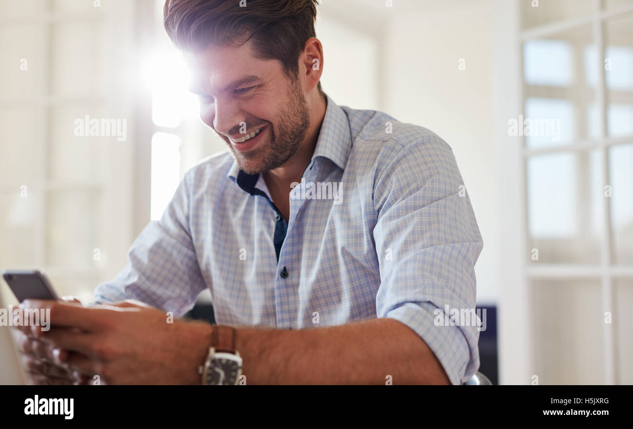 Indoor shot of happy young man sitting at home using mobile phone. Handsome caucasian male reading text message on his smart pho Stock Photo