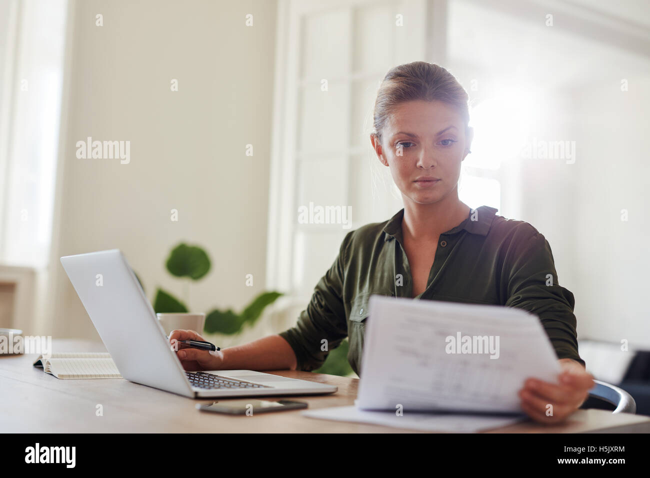 Shot of young woman sitting at table with laptop and reading documents. Beautiful businesswoman working from home office. Stock Photo