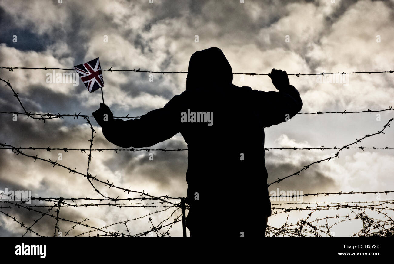 Rear view of man wearing hooded jacket holding Union Jack flag looking through razor wire fence. Brexit, immigration... concept Stock Photo