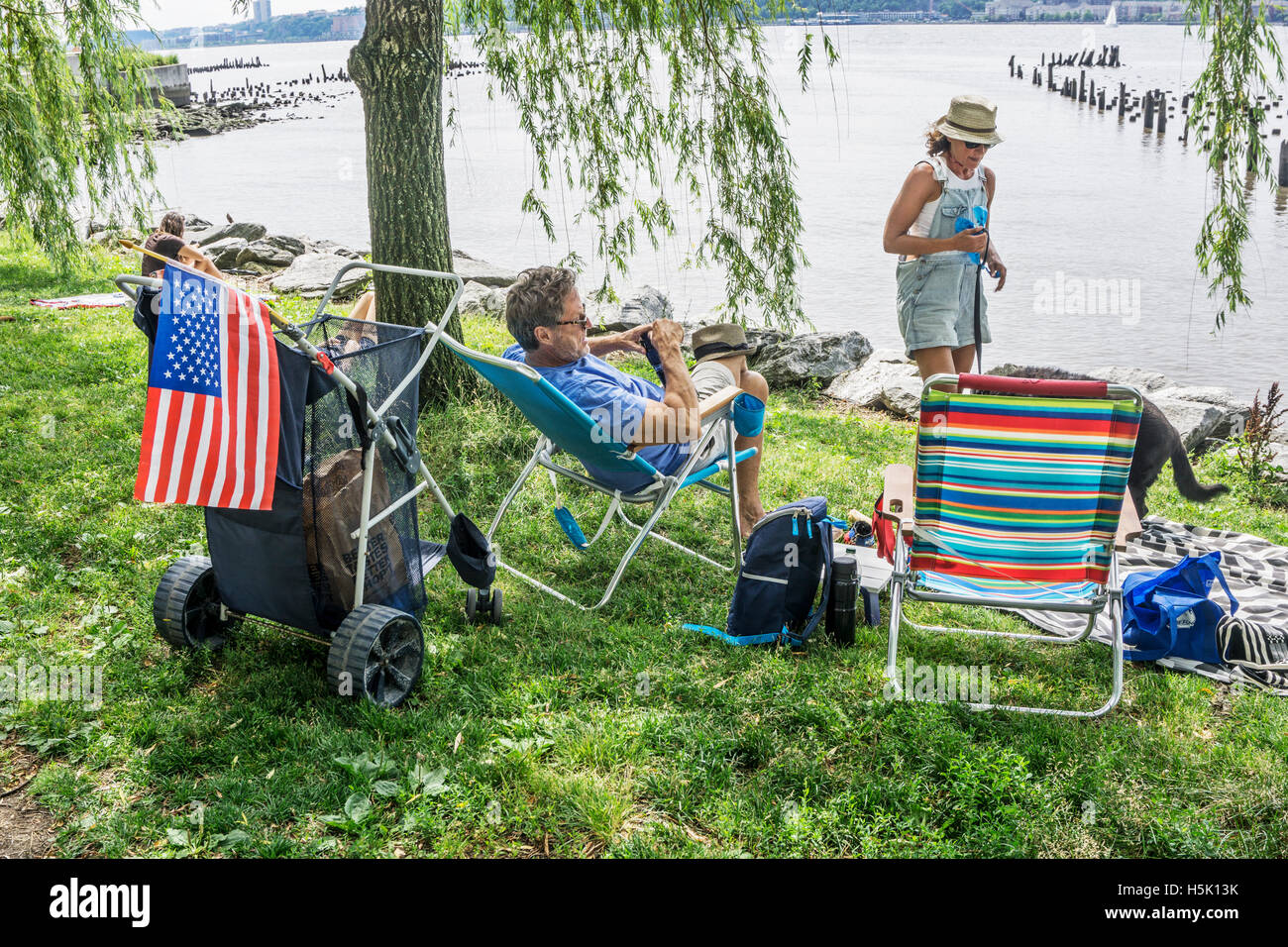 couple with American flag taped to loaded shopping cart prepare to picnic on lawn at edge of water to celebrate July 4th holiday Stock Photo