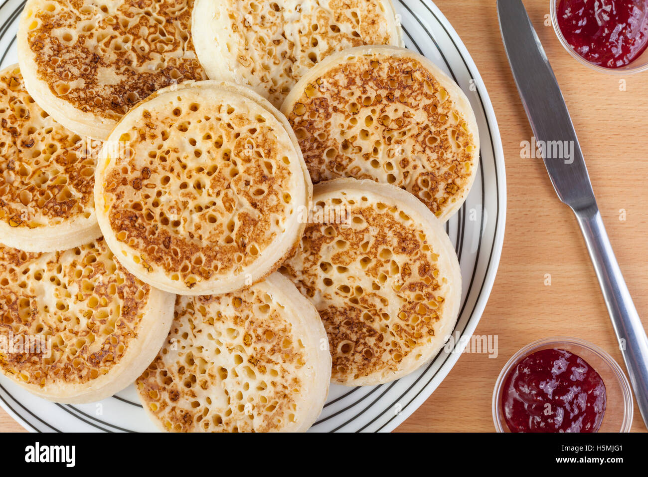 Large plate of freshly toasted crumpets and strawberry jam with a knife on a breakfast table Stock Photo