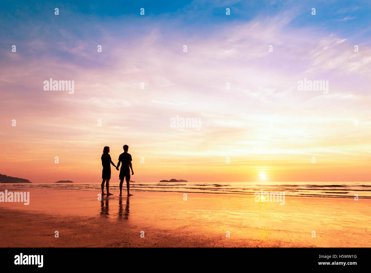 Two lovers standing together on a tropical beach and thinking about life, beautiful sunset in the background Stock Photo