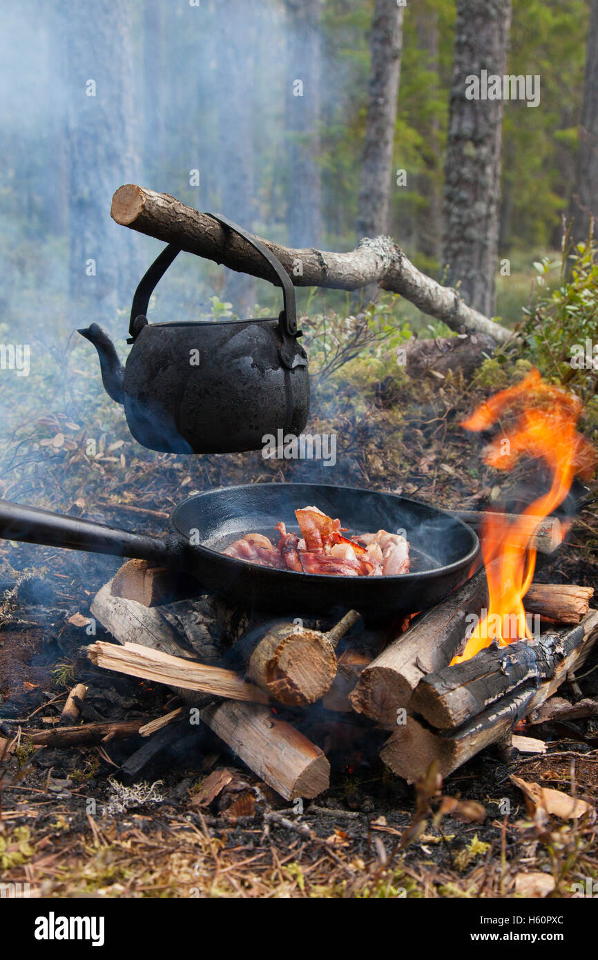 Blackened tin kettle boiling water and pan cooking bacon over flames from campfire during hike in forest Stock Photo