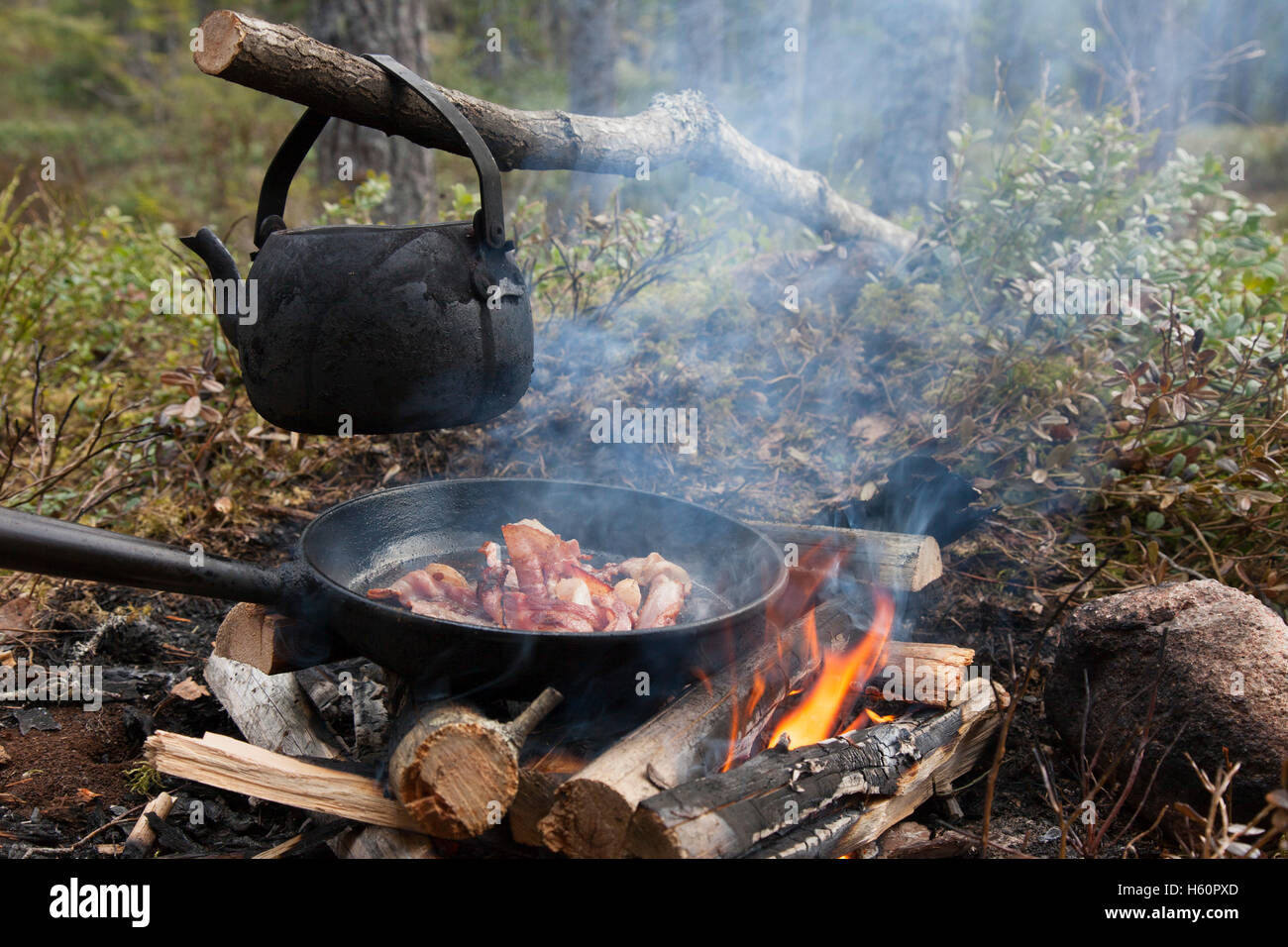 Blackened tin kettle boiling water and pan cooking bacon over flames from campfire during hike in forest Stock Photo