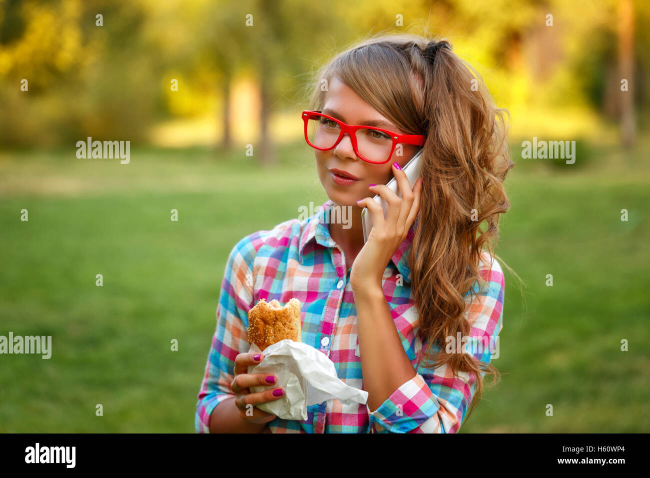 Pretty attractive girl talking on the phone and holding a hot dog in a park. Snack. Fast food. Stock Photo