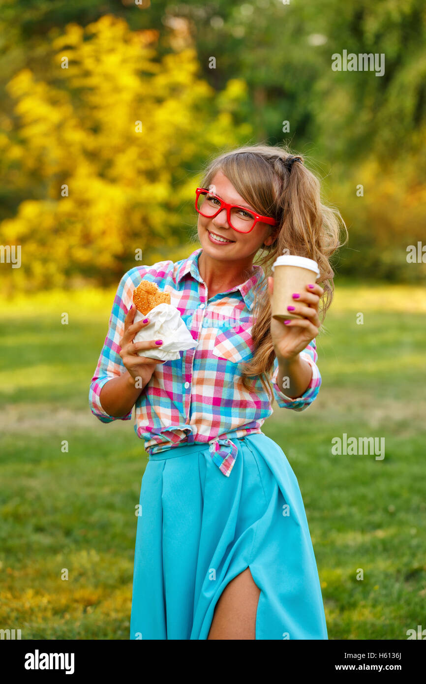 Pretty attractive girl holds cup of coffee and a hot dog in a park. Snack. Fast food. Student at dinner. Stock Photo