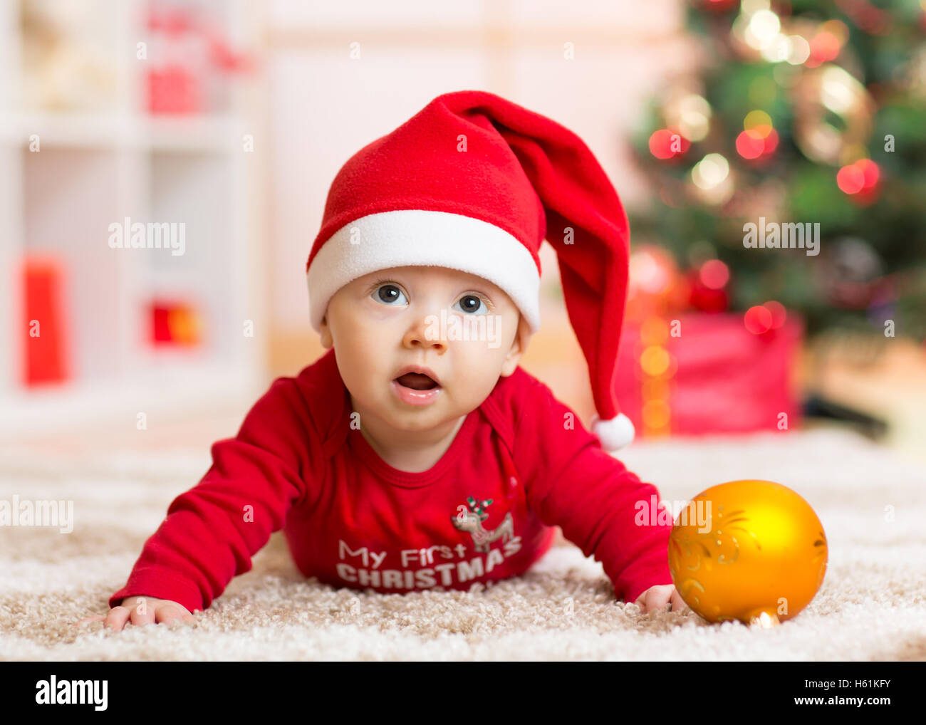 Funny baby lying on tummy wearing Santa hat and suit on floor in front of Christmas tree Stock Photo
