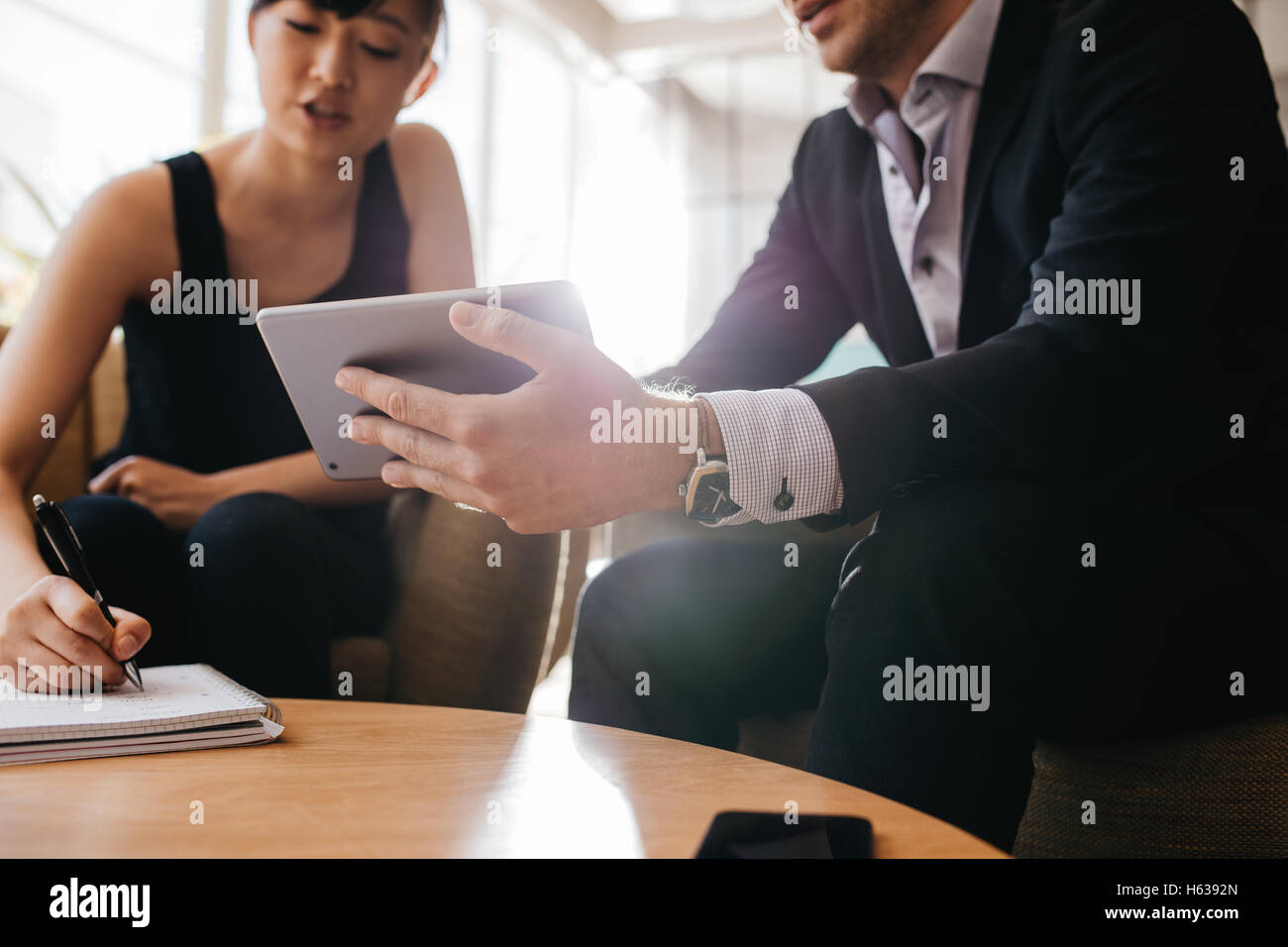 Cropped image of businessman holding digital tablet and woman with notepad. Businesspeople sitting in lobby working together. Stock Photo
