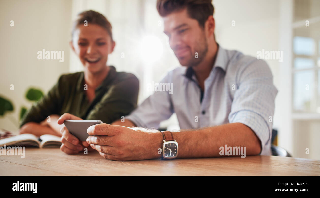 Shot of young man sitting with his girlfriend at table holding smart phone. Young couple at home using mobile phone. Focus on ma Stock Photo