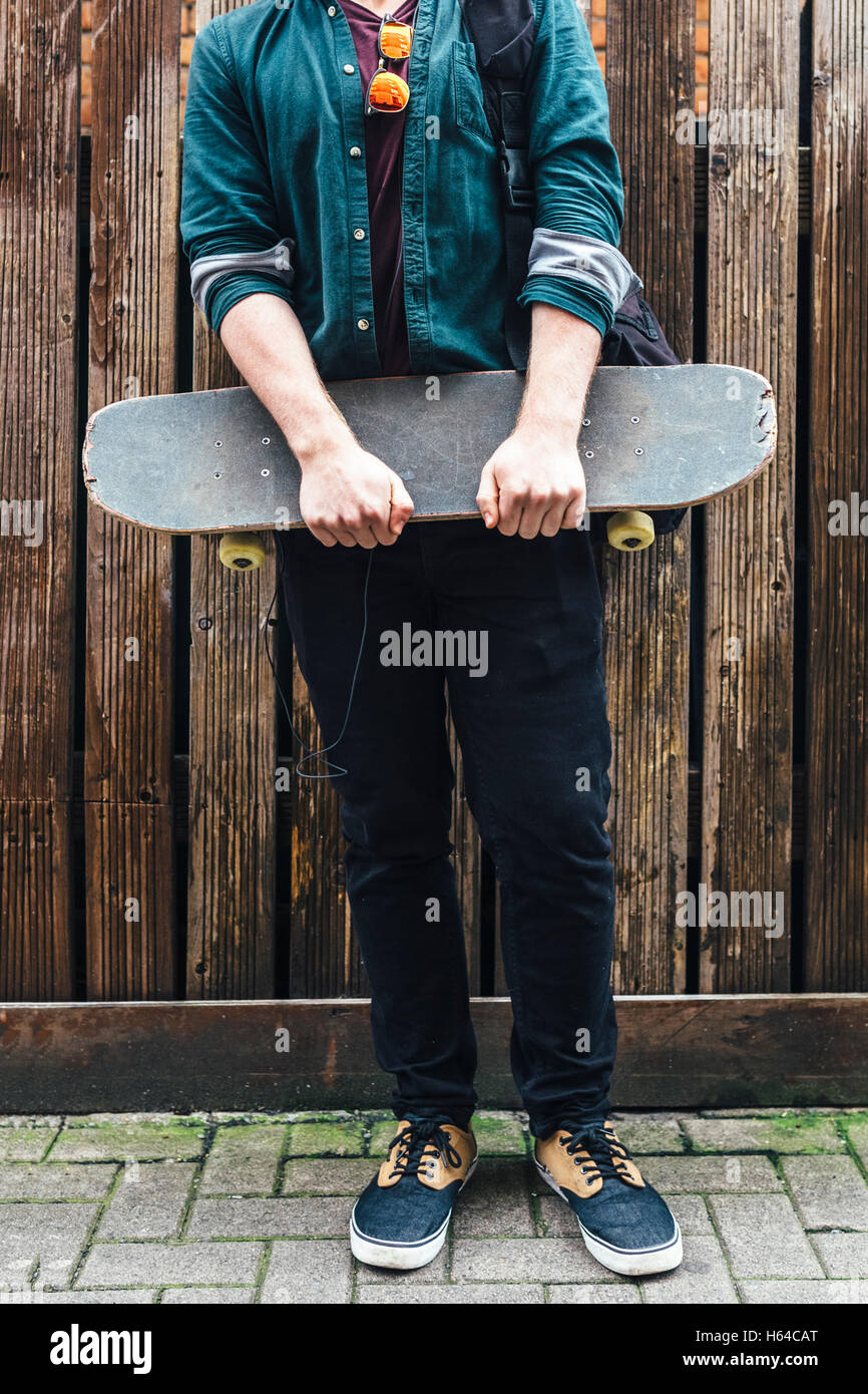 Young man standing on pavement holding skateboard, partial view Stock