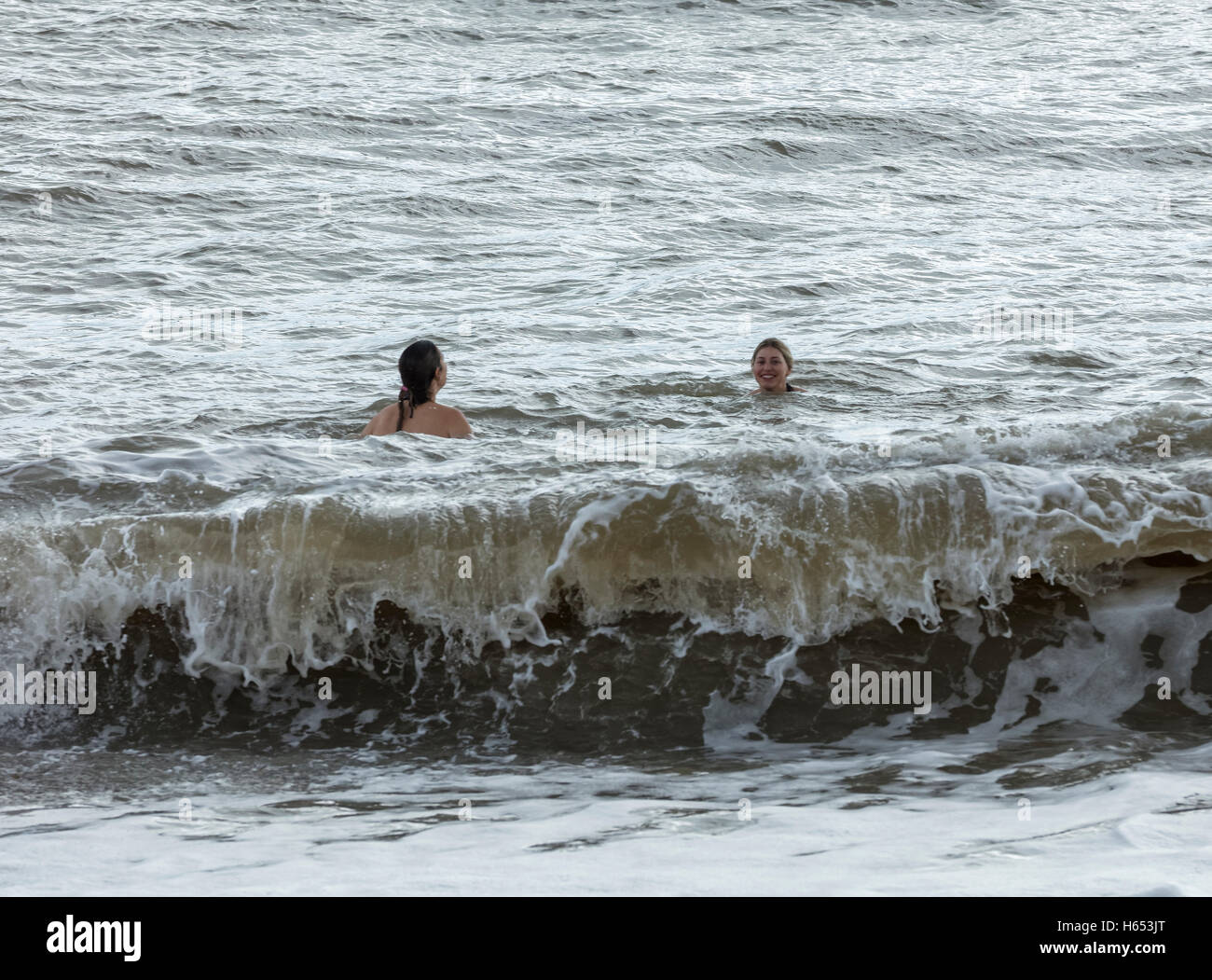 Two young women swmming in a very cold sea at Aldeburgh in Suffolk with a large wave. One of the women has her back to camera Stock Photo