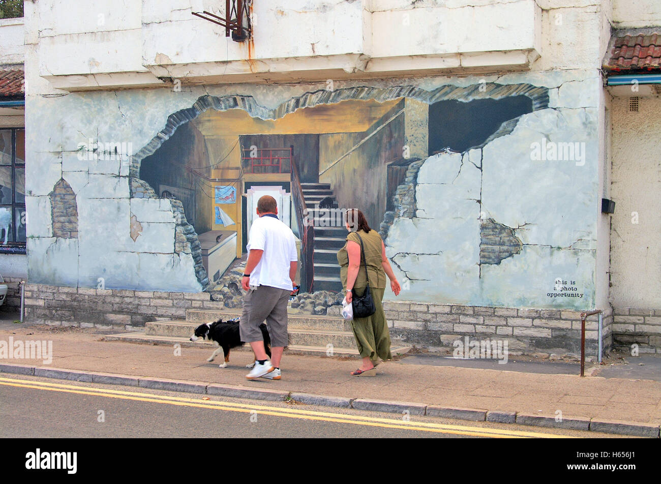 Couple walking past Trompe l'oeil effect Mural art painting on the side of a building in Swanage town, Dorset, England. Stock Photo